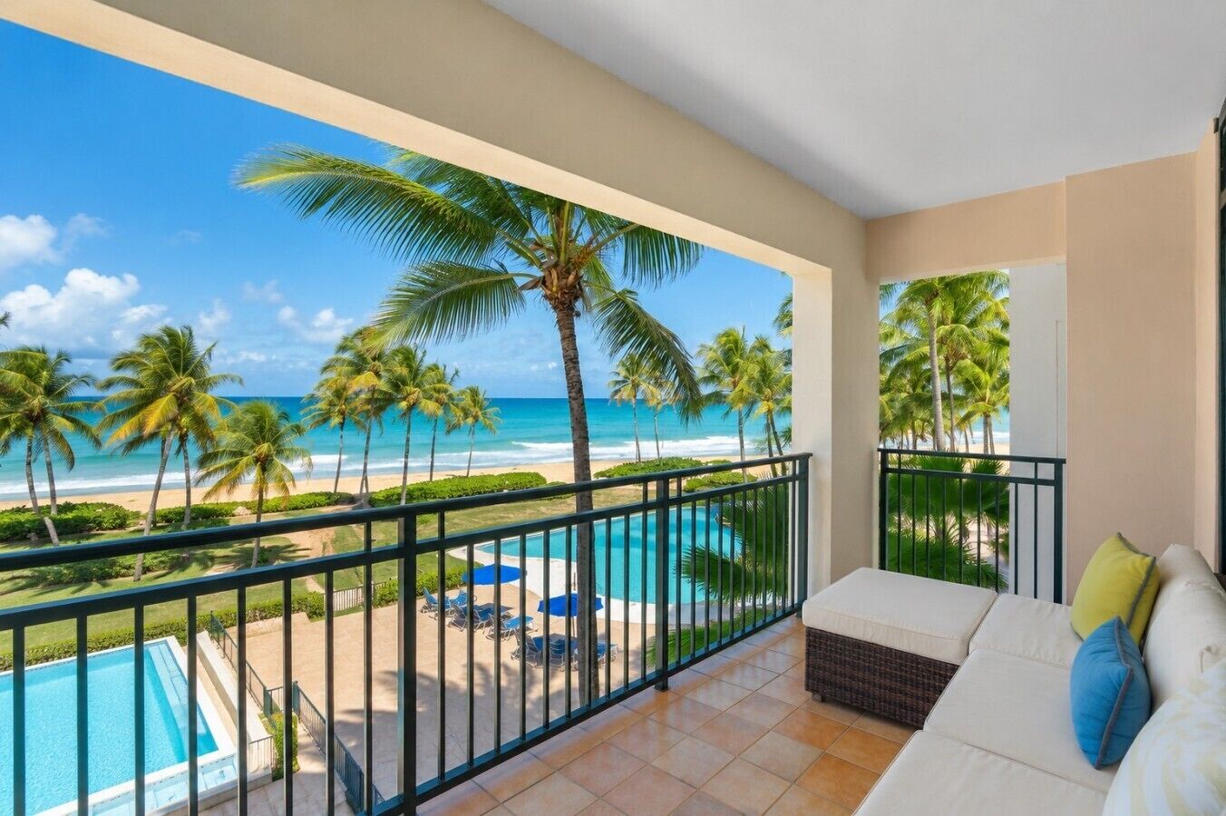 A balcony with a view of the beach and ocean, a pool, and a seating area with cushions.