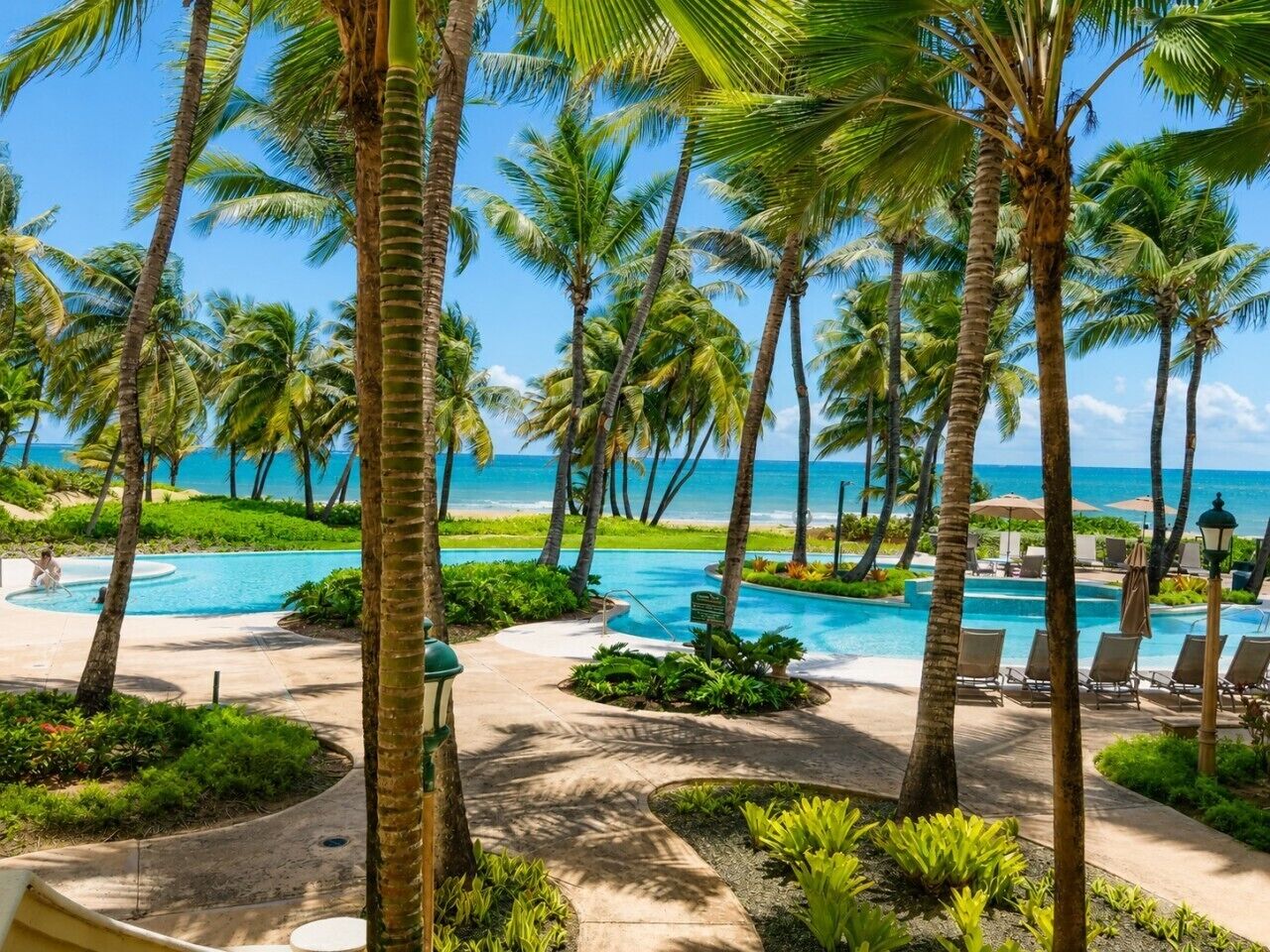Ein Poolbereich eines Resorts mit Palmen, Liegestühlen und Blick auf den Ozean.