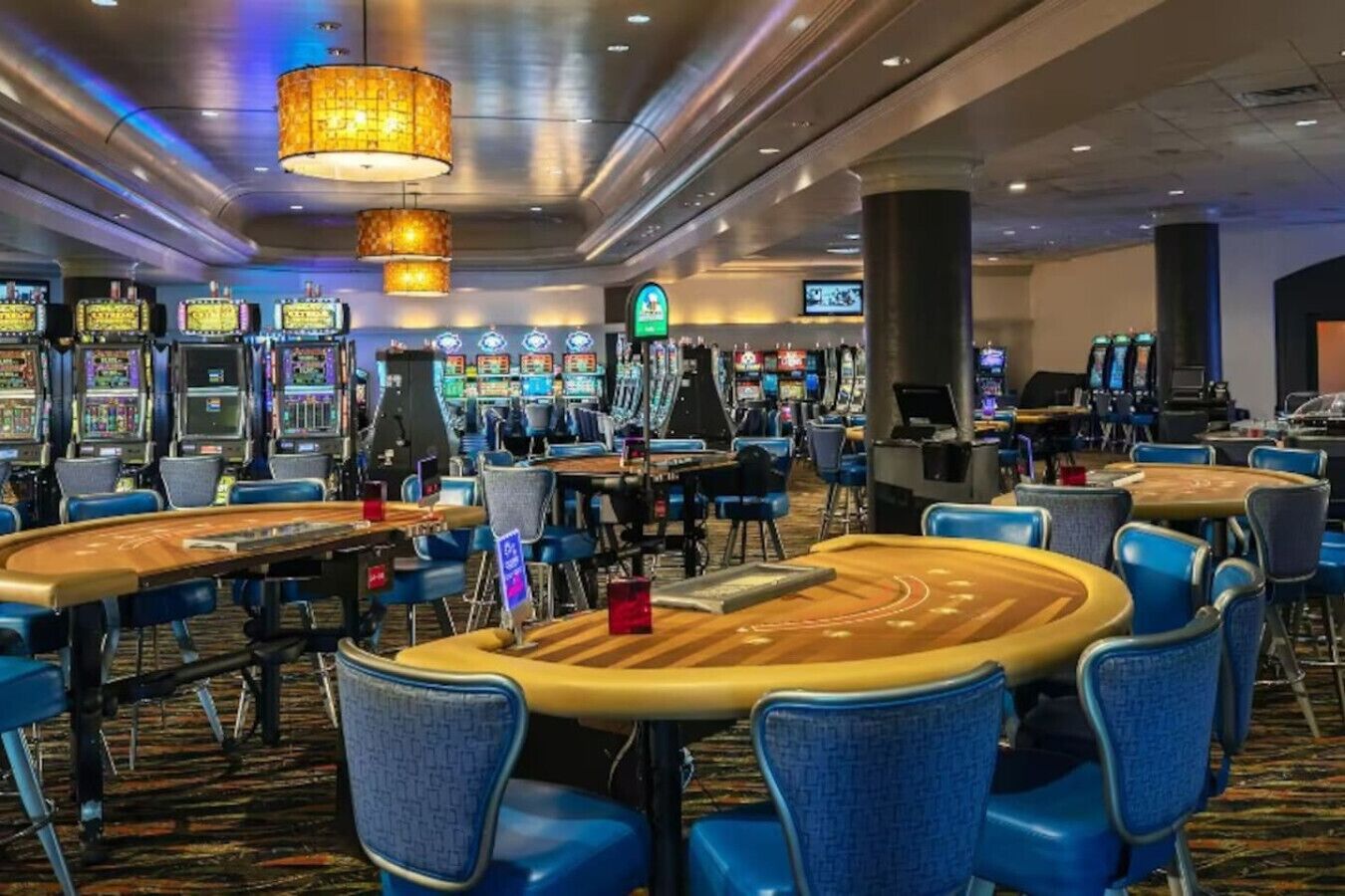 A casino with multiple gaming tables and chairs, illuminated by hanging lamps.