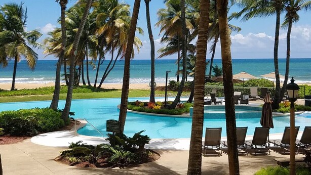 Zona de piscina en un resort, con palmeras, sillas de descanso y vista al océano.