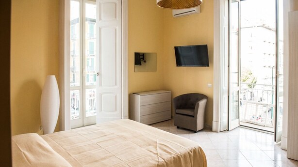 Un dormitorio con cama, cómoda, silla, televisor y balcón con vista a edificios.
