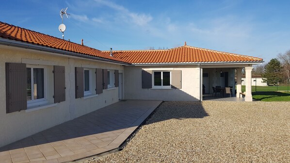 A building with a tiled roof, multiple windows with shutters, a gravel driveway, and a covered outdoor seating area.