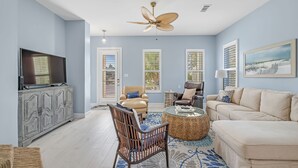 A living room with a flat-screen TV, wicker chairs, a beige sofa, and a ceiling fan. - Avalon - 3BR/3BA beach house with beach view (Gulf Shores)