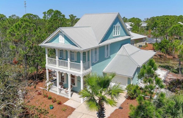 A two-story house with a balcony, surrounded by trees and a sandy area.