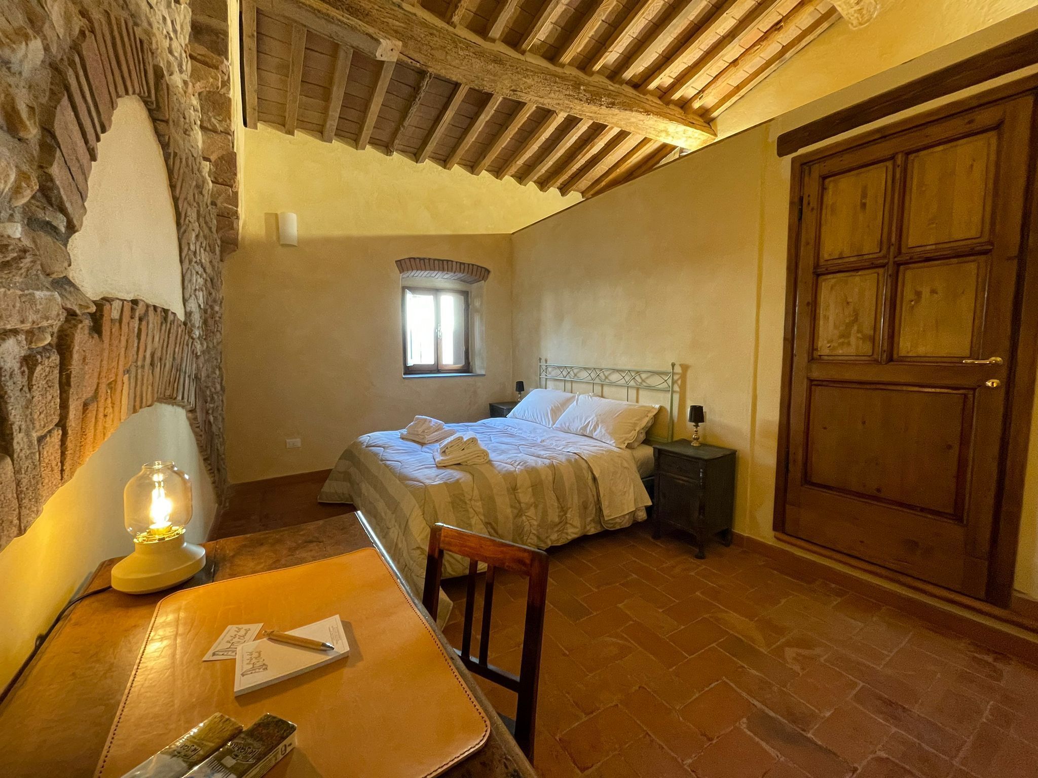 Ein Schlafzimmer mit Steinsandsteinwand, Holzbalkendecke, einem Bett mit weißen Leinentüchern, einem Nachttisch mit Lampe, einer Holztür und einem Fenster mit Tageslicht.