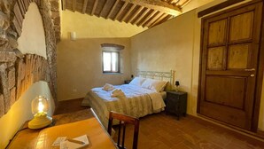 A bedroom with a stone wall, a wooden ceiling, a bed with white linens, a nightstand with a lamp, a wooden door, and a window with natural light. - Characteristic apartment in historic 1300s tower (San Casciano in Val di Pesa)