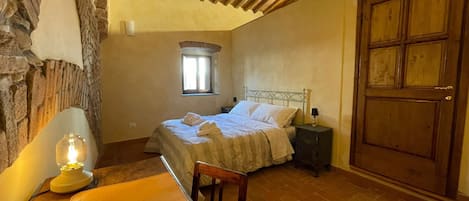 Un dormitorio con pared de piedra, techo de madera, cama con ropa blanca, mesita de noche con lámpara, puerta de madera y ventana con luz natural.