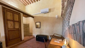 A room with a wooden door, a small table with a lamp, a suitcase, and a wall-mounted air conditioner. - Characteristic apartment in historic 1300s tower (San Casciano in Val di Pesa)