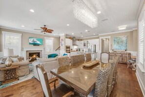 A spacious living room with a fireplace, a dining area with a wooden table and woven chairs, and a kitchen with white cabinets and stainless steel appliances.