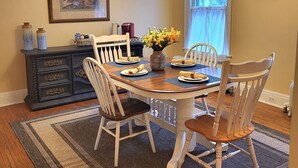 A dining room with a wooden table set for four, surrounded by white chairs. A dark wooden dresser with drawers and a vase of flowers is against the wall. - Huge deck & yard Near Indoor Sports Arena! Spacious! Smart TVs! Fast Wifi! Cozy. (Machesney Park)