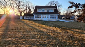 A two-story house with a porch, surrounded by a grassy yard and trees. - Huge deck & yard Near Indoor Sports Arena! Spacious! Smart TVs! Fast Wifi! Cozy. (Machesney Park)