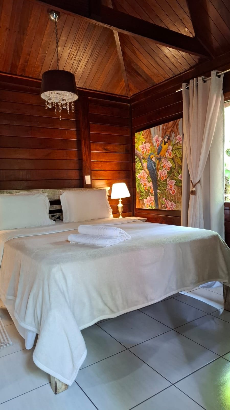 A hotel room with a wooden ceiling, a bed with white linens, a nightstand with a lamp, and a painting of birds on the wall.