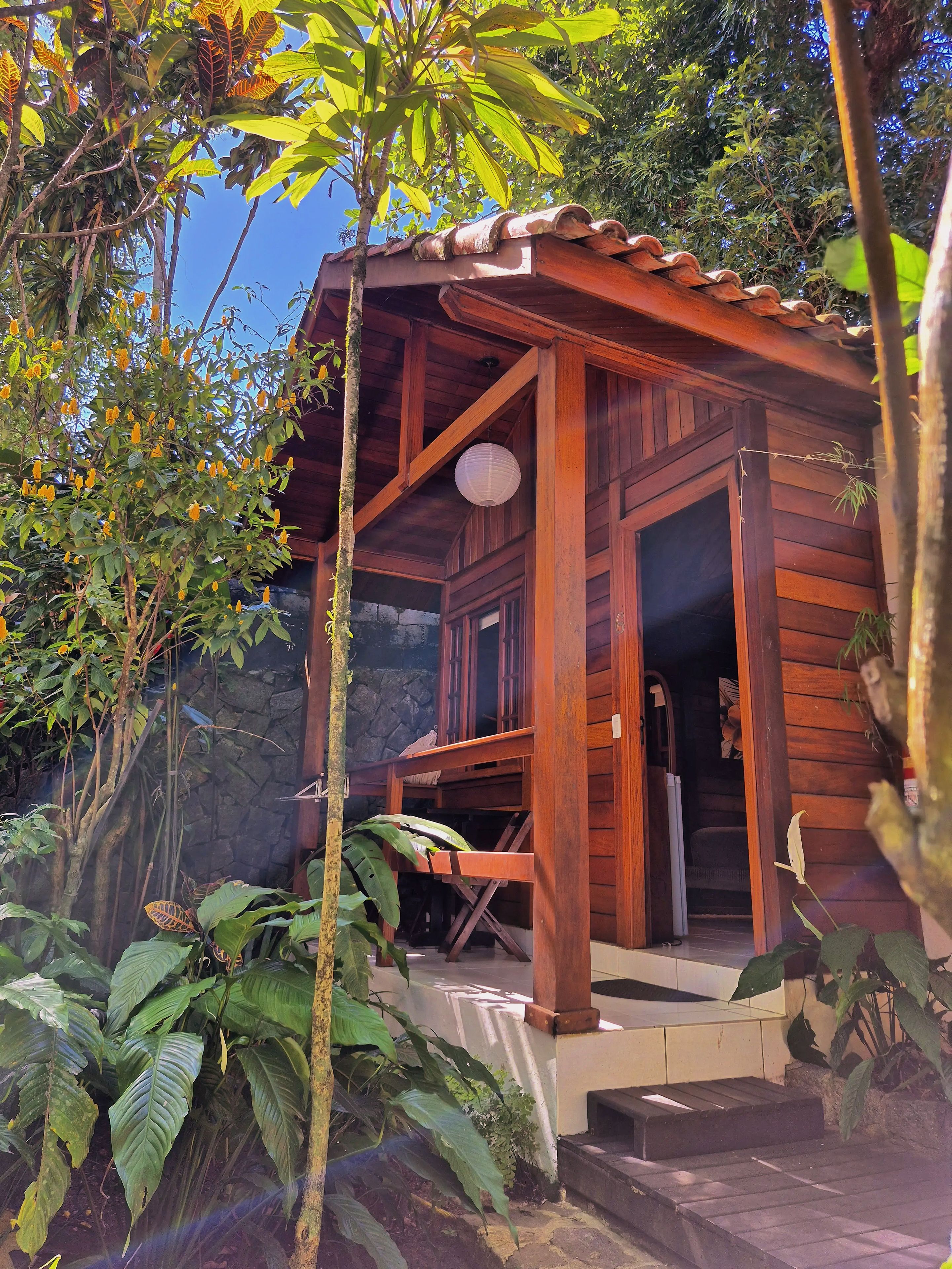 Una cabaña de madera con porche, rodeada de abundante vegetación y un muro de piedra.