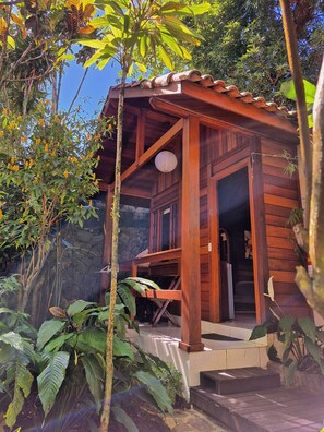 A wooden cabin with a porch, surrounded by lush greenery and a stone wall. - Aratinga inn (Angra dos Reis)