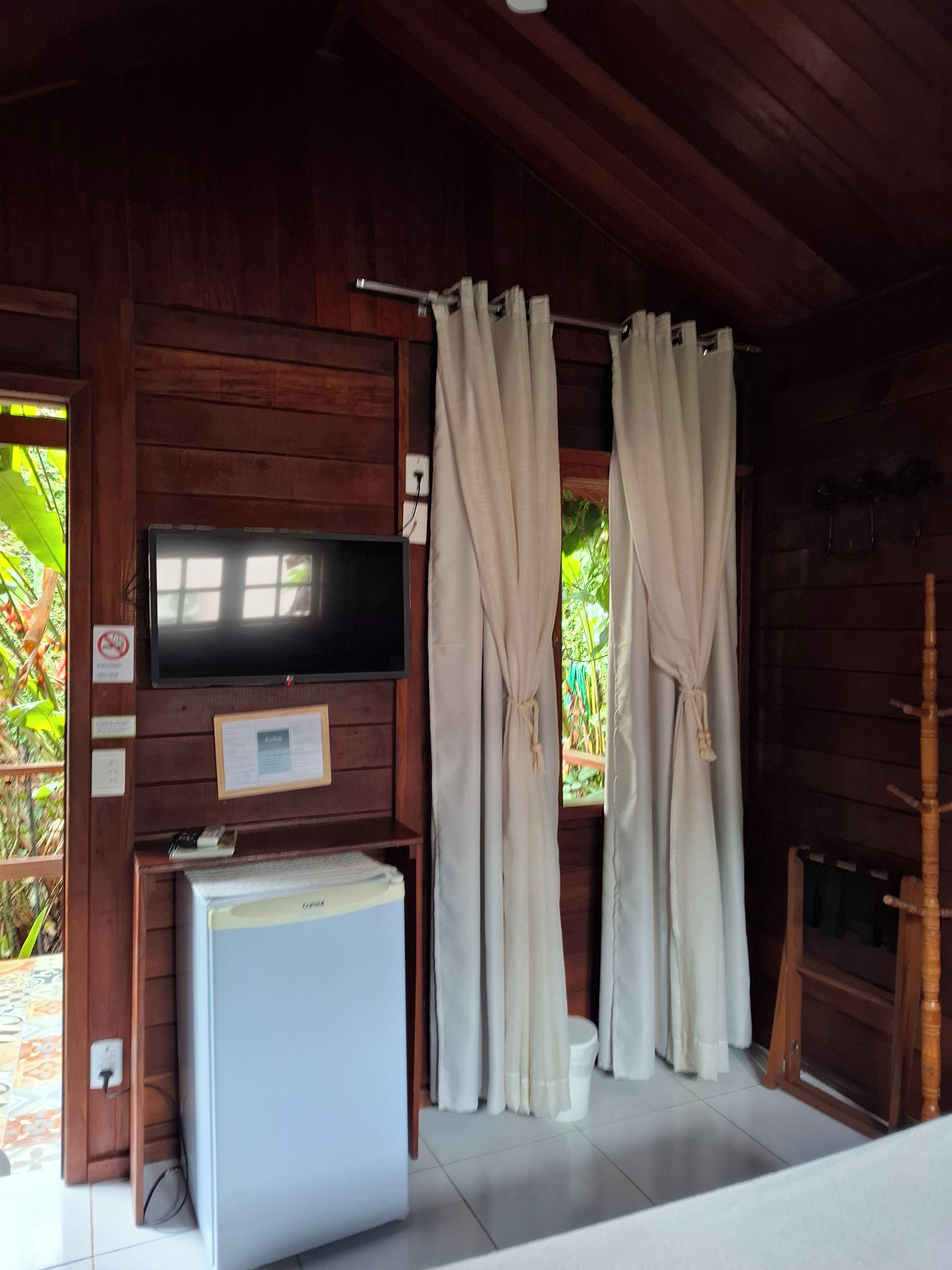 Una habitación con televisión instalada en la pared, un refrigerador pequeño y cortinas en las ventanas.