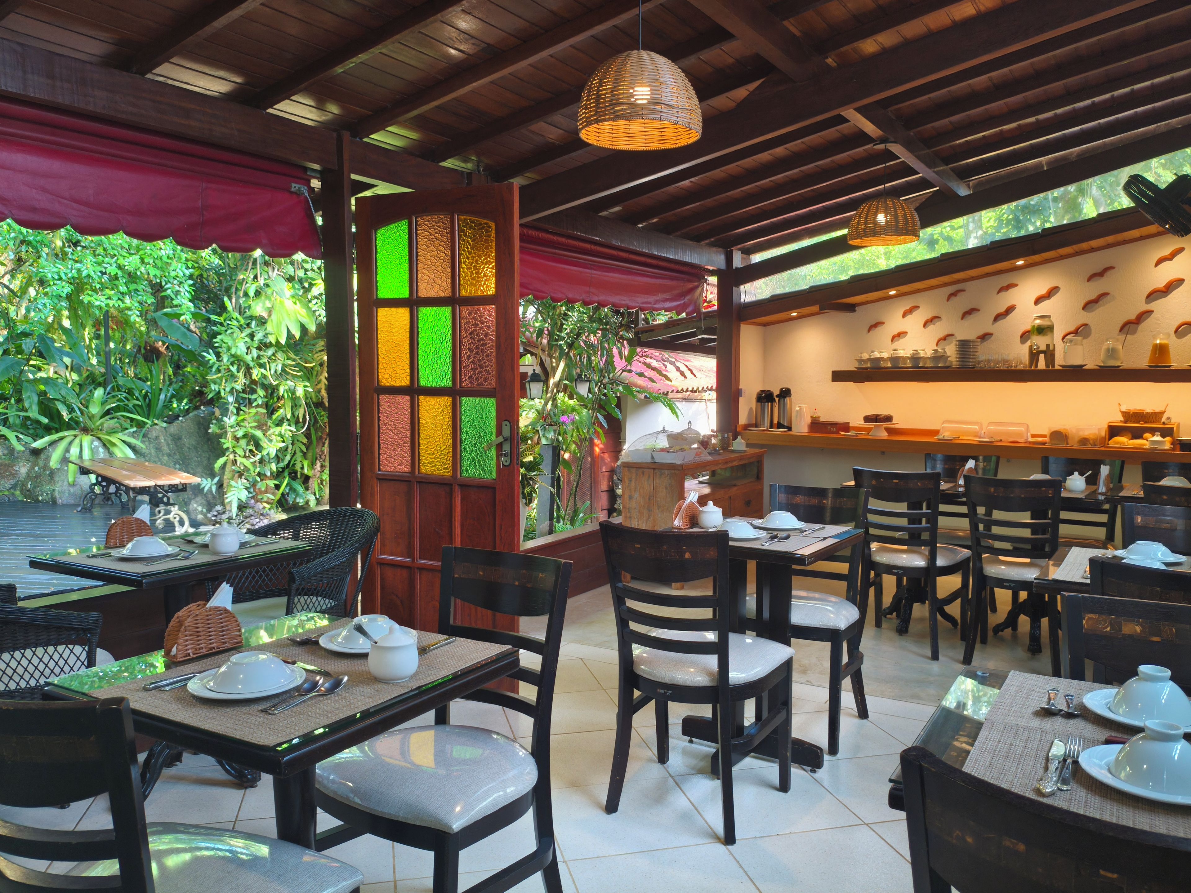 An outdoor dining area with tables and chairs, a wooden ceiling, and a view of greenery.