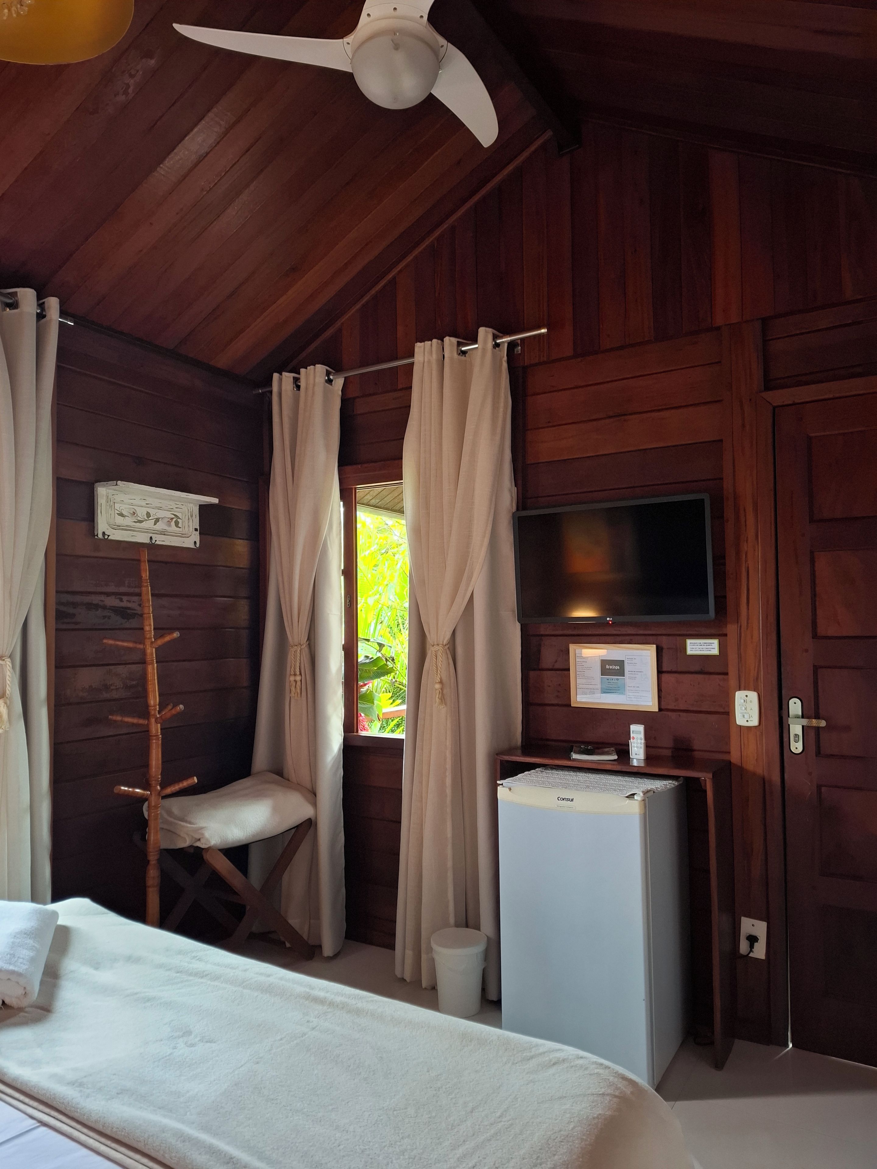 A room with wooden walls, a ceiling fan, a bed, a small table, a television, and a window with curtains.