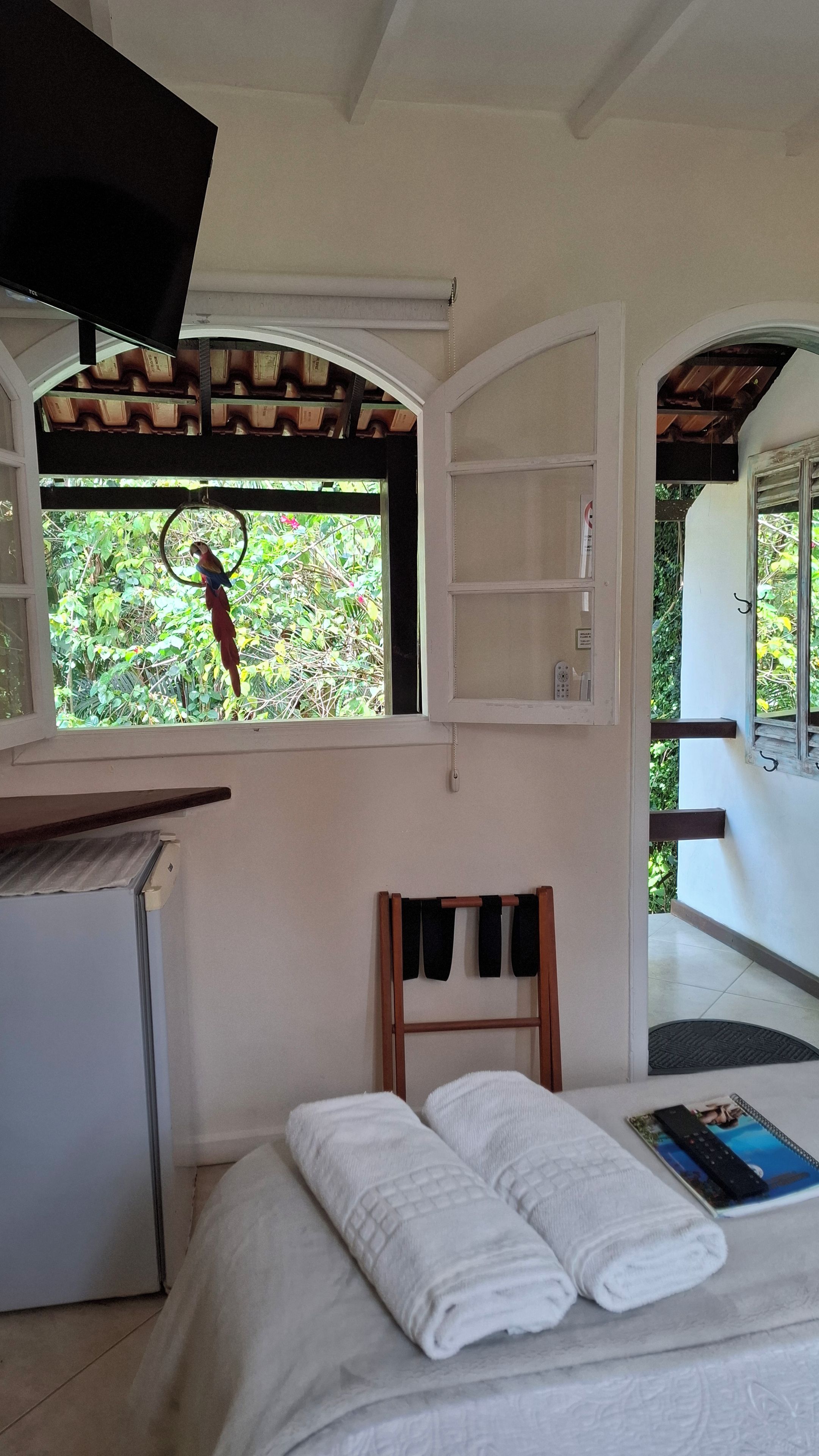 A hotel room with a bed, a desk, a window with a view of greenery, and a door leading outside.
