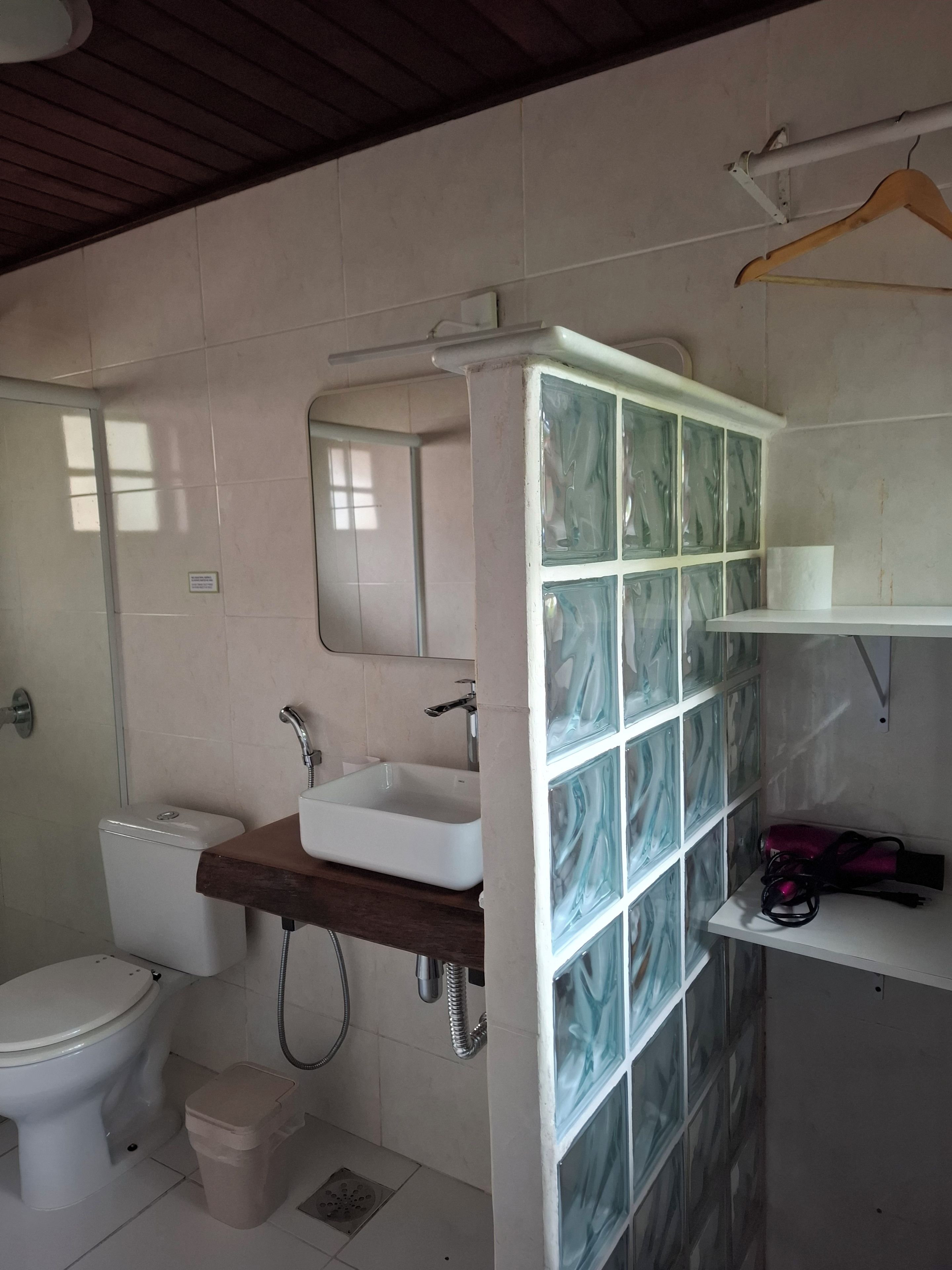 A bathroom with a toilet, sink, mirror, shower enclosure, and a shelf with a bag.
