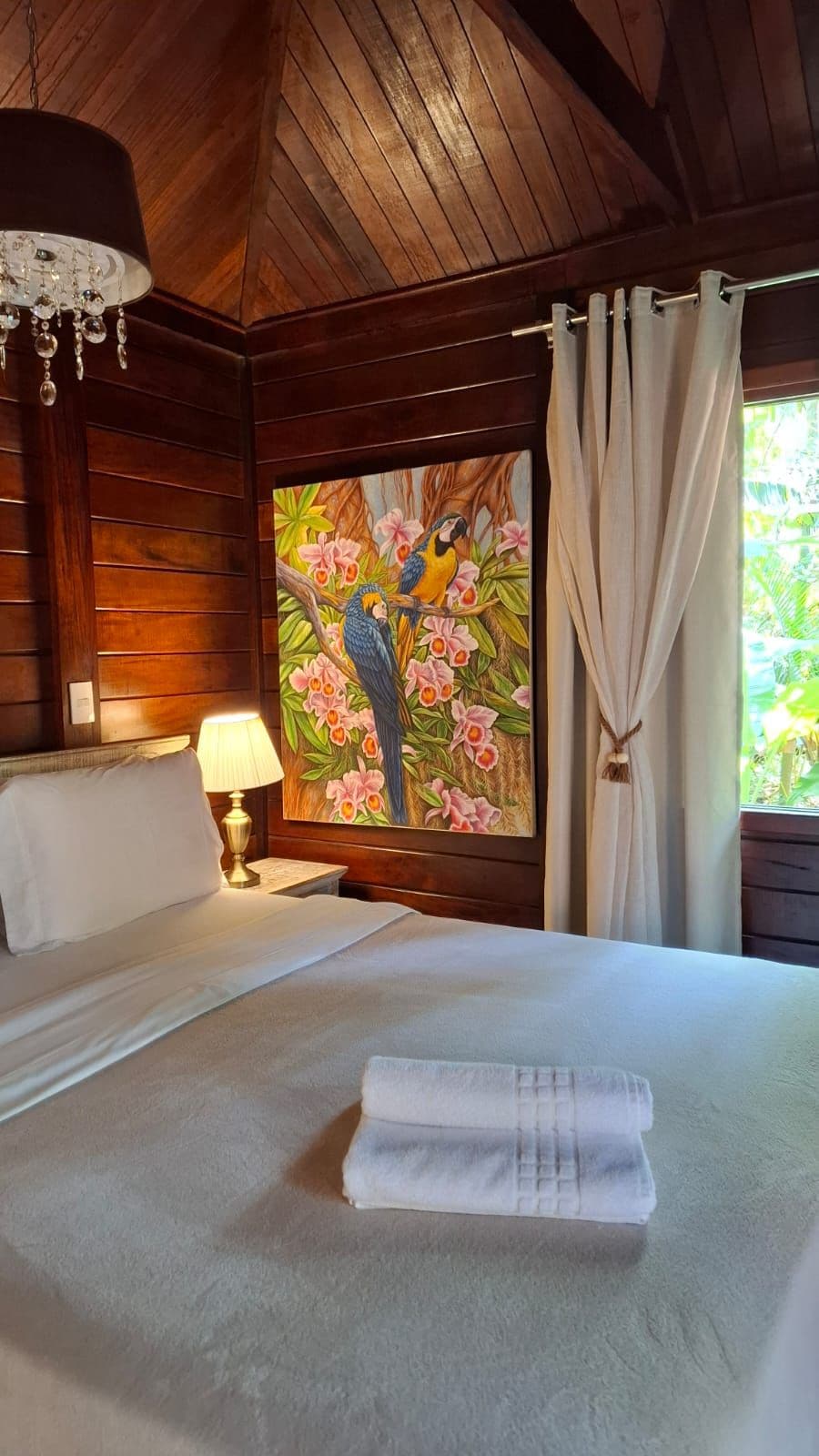 A hotel room with a bed, a painting of parrots, a lamp, and curtains.