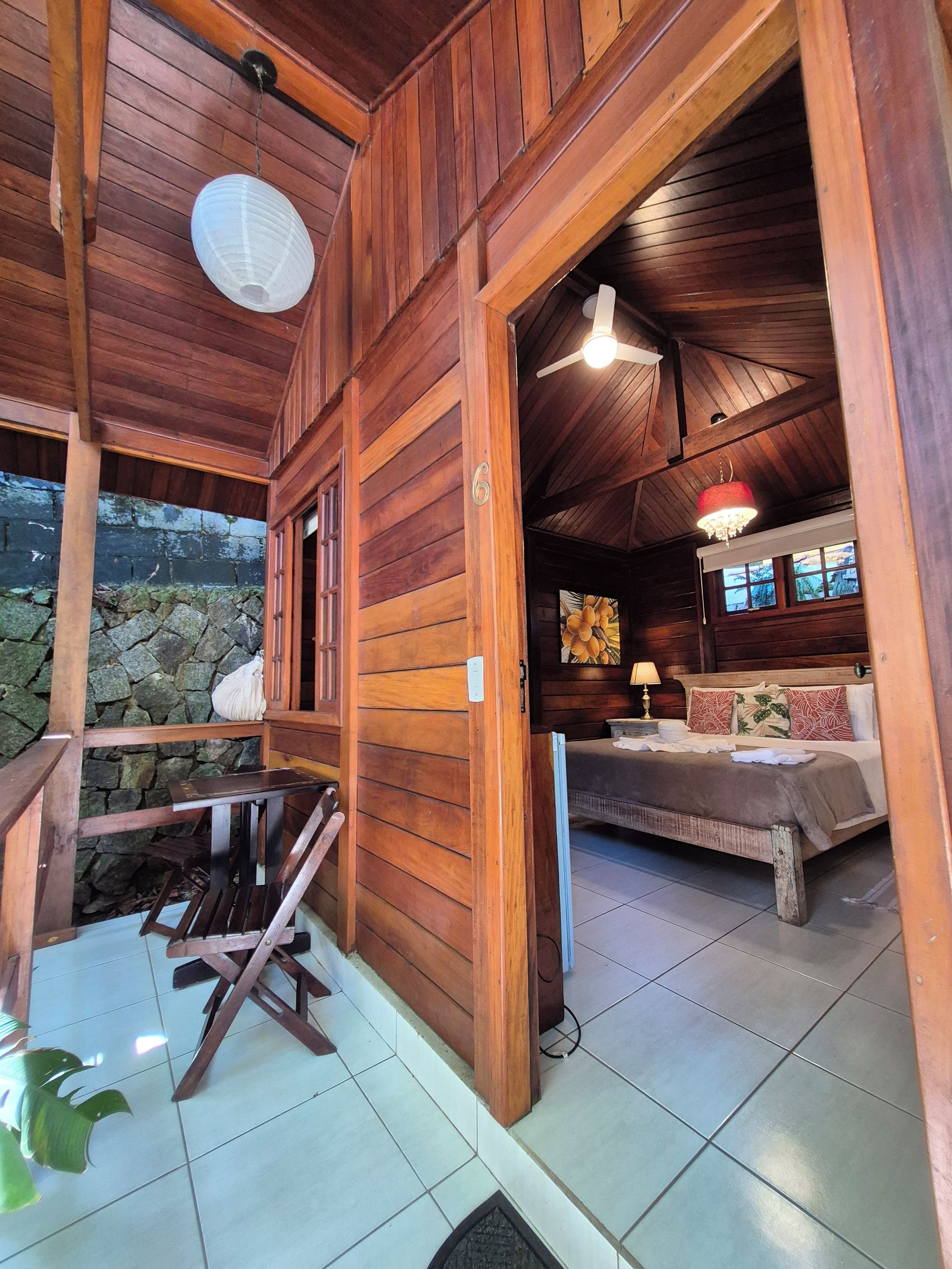 A wooden cabin interior with a bed, a hanging light fixture, and a stone wall outside.