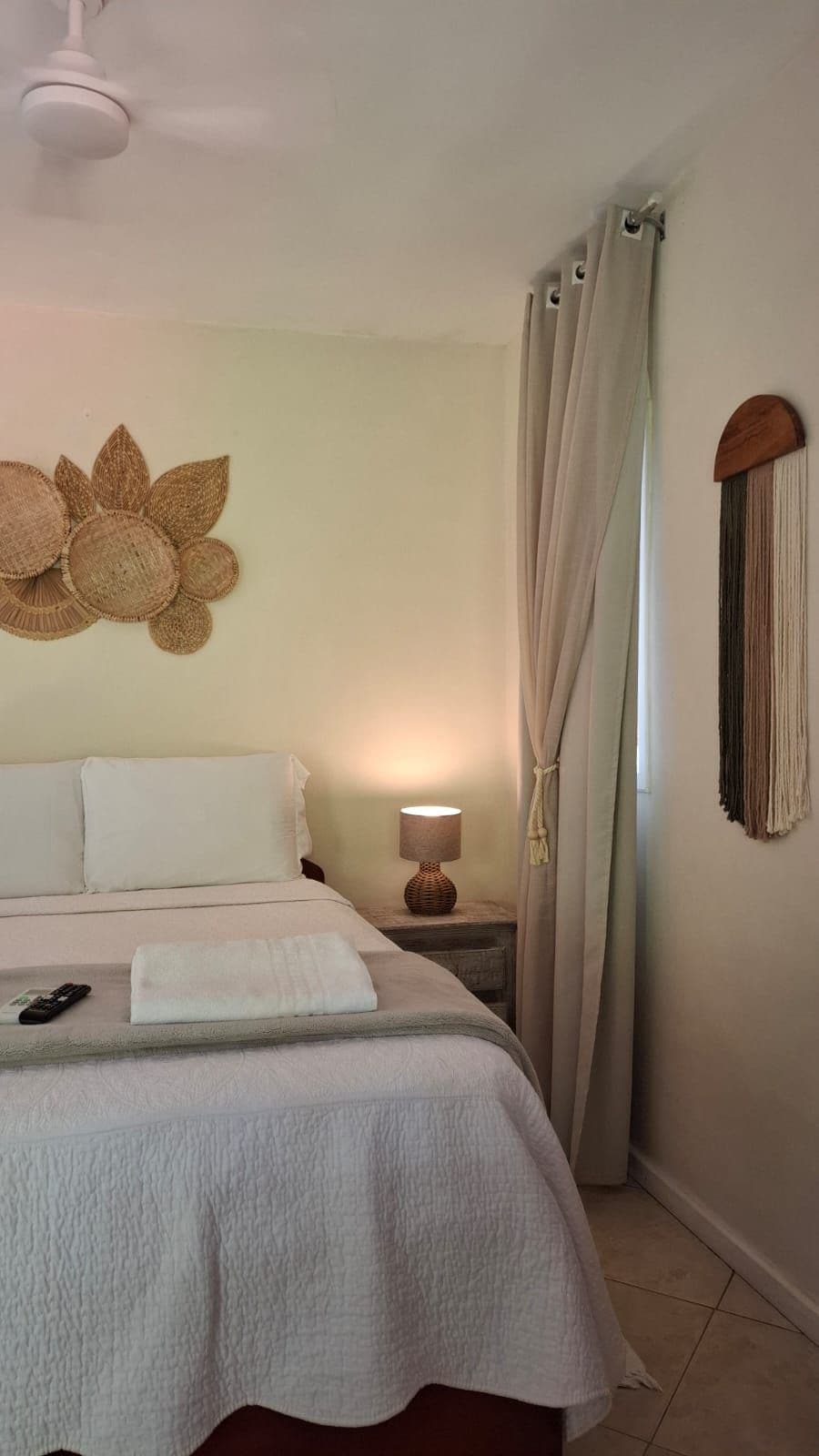 Quarto de hotel com cama, mesa de cabeceira com luminária, objeto decorativo na parede e cortina.