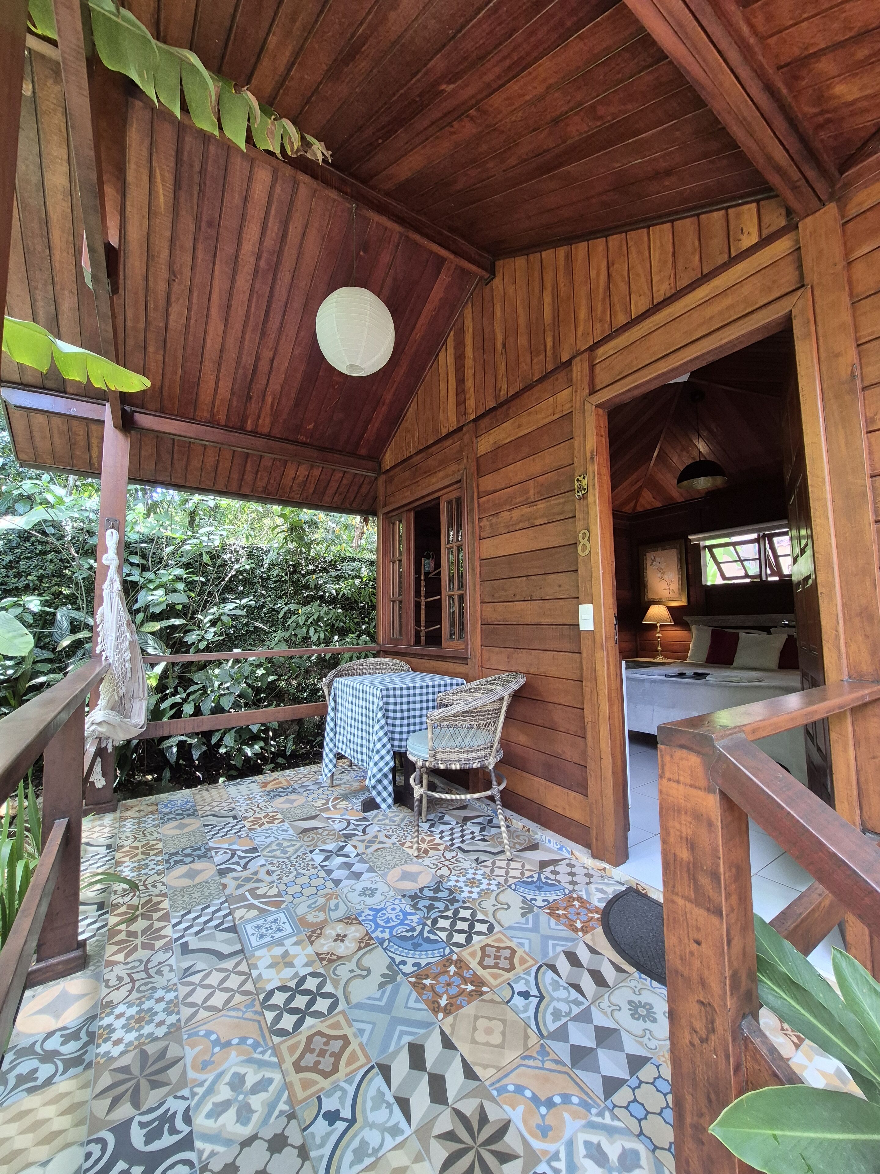 Une cabane en bois avec une table à carreaux et une chaise en osier sur une véranda carrelée, qui donne sur une chambre à coucher avec un lit et une table de chevet.