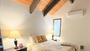 Un dormitorio con una cama, dos mesitas de noche, una lámpara, un ventanal y un techo con vigas a la vista.
