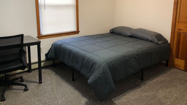 Una habitación pequeña y poco amoblada, con una cama individual, un escritorio y una ventana con persianas.