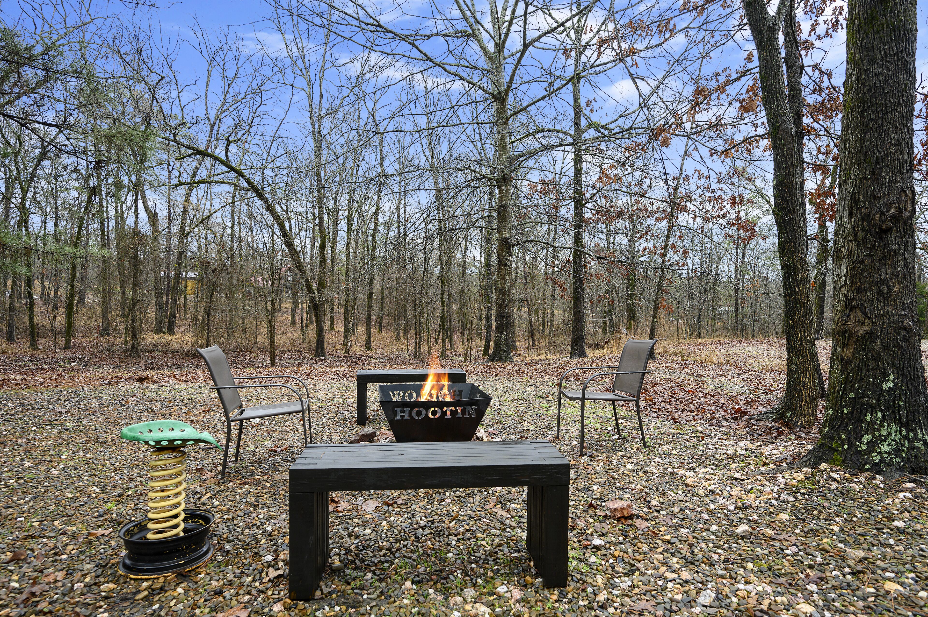 Un espace extérieur aménagé avec un foyer, deux chaises et une table, le tout situé dans un cadre forestier.