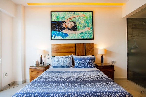 Un dormitorio con una cama, dos mesitas de noche, un velador, un cuadro en la pared y una puerta.