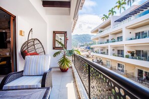 A balcony with a wicker chair, a potted plant, and a view of a cityscape with buildings and palm trees.