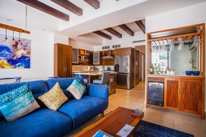 A modern living room with a blue sofa, wooden cabinets, and a kitchen area with a refrigerator and stove. - Workspace, Mini-Bar & Rooftop Pool | 2BR Retreat (Puerto Vallarta)