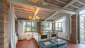 Quarto com cama, um banquinho, mesa de cabeceira e janela com cortinas.
