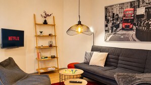 A living room with a grey sofa, a red chair, a wooden bookshelf, a round coffee table, a hanging light fixture, and a TV displaying 'NETFLIX'.