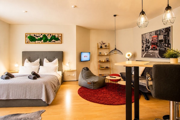 A modern hotel room with a bed, a desk, a chair, a red rug, a bookshelf, and a wall-mounted TV.