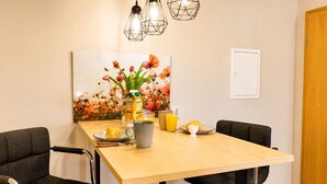 A dining area with a wooden table, black chairs, a painting of flowers, a fruit bowl, a juice carton, and a coffee mug.