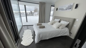 Ein modernes Schlafzimmer mit einem großen Bett, einem weißen Teppich und Blick auf eine verschneite Landschaft durch große Fenster.