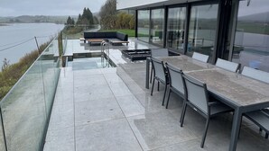 Eine moderne Terrassenanlage im Freien mit Glasbrüstung und Blick auf ein Gewässer, ausgestattet mit einem langen Tisch, Stühlen und einer schwarzen Couch.