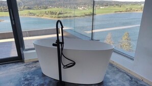 Ein modernes Badezimmer mit freistehender Badewanne, großen Fenstern und Blick auf einen See und Hügel.