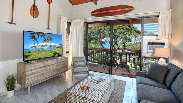 A modern living room with a TV, wooden furniture, a balcony with a view of the sea, and a ceiling with wooden beams.