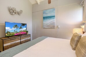 A bedroom with a bed, a TV on a wooden dresser, a painting of a beach, and a lamp.