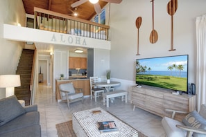 A spacious living area with a high ceiling, a dining set, a TV displaying a tropical scene, and a staircase leading to an upper level. - The Ultimate Escape. Relax, Enjoy, Steps to Beach (Kihei)