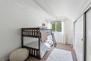 Un dormitorio con una cama litera, un reposapiés redondo pequeño, una alfombra blanca y una ventana con cortinas.
