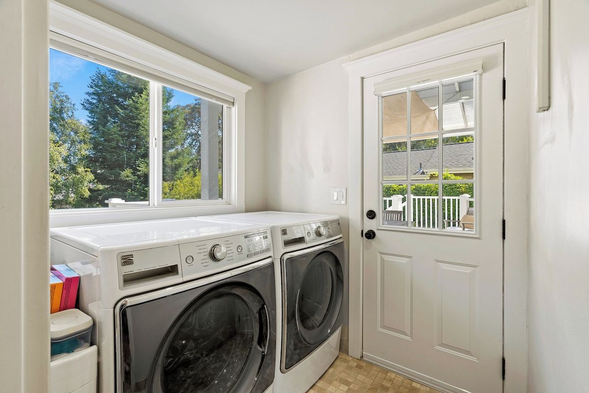A laundry room with two front-loading washing machines, a window with a view of trees, and a white door.