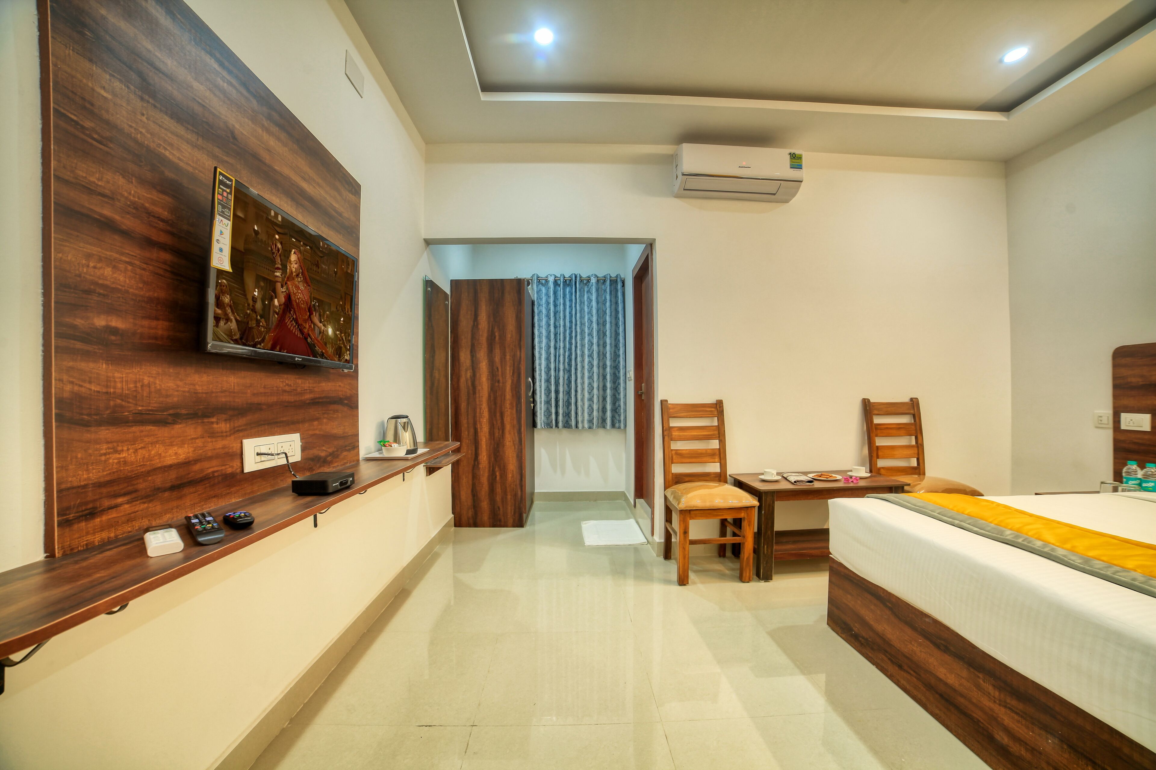 A hotel room with a wooden accent wall, a bed with a yellow bedspread, a desk with a chair, a television mounted on the wall, and an air conditioning unit on the ceiling.