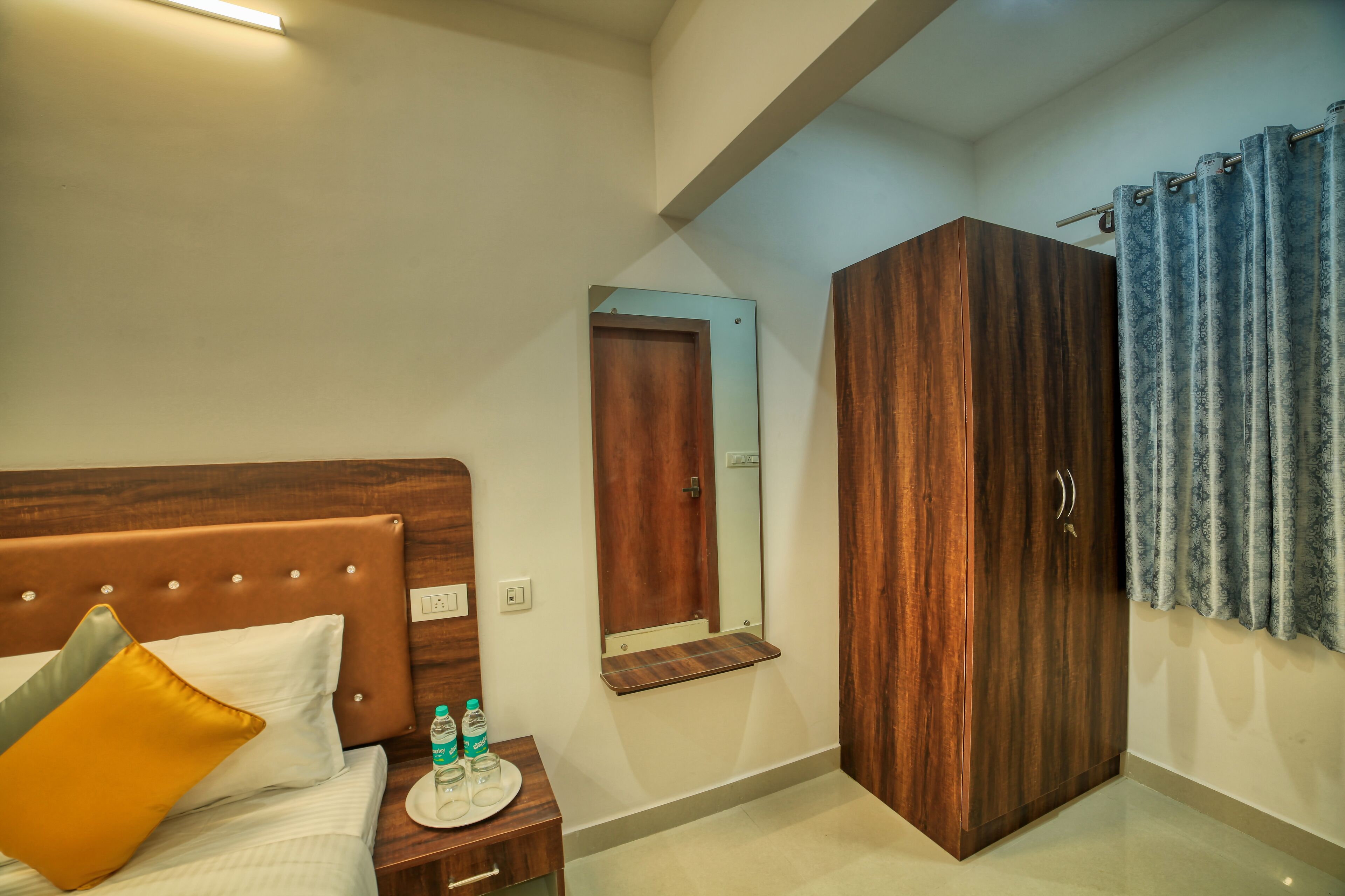 Habitación de hotel con cabecera de madera, cama con almohadas blancas y amarillas, mesita de noche con botella de agua, armario de madera y espejo.