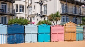 A row of colorful beach huts with a sandy beach in front and a building with balconies in the background. - Liberty Cottage, Broadstairs (Broadstairs)