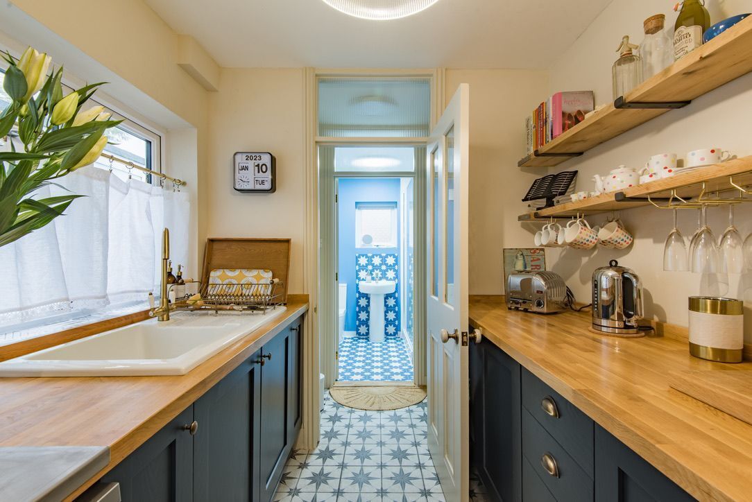 A narrow kitchen with a sink, wooden countertop, and a blue and white tiled floor. There is a door leading to another room, and shelves with kitchenware and books.