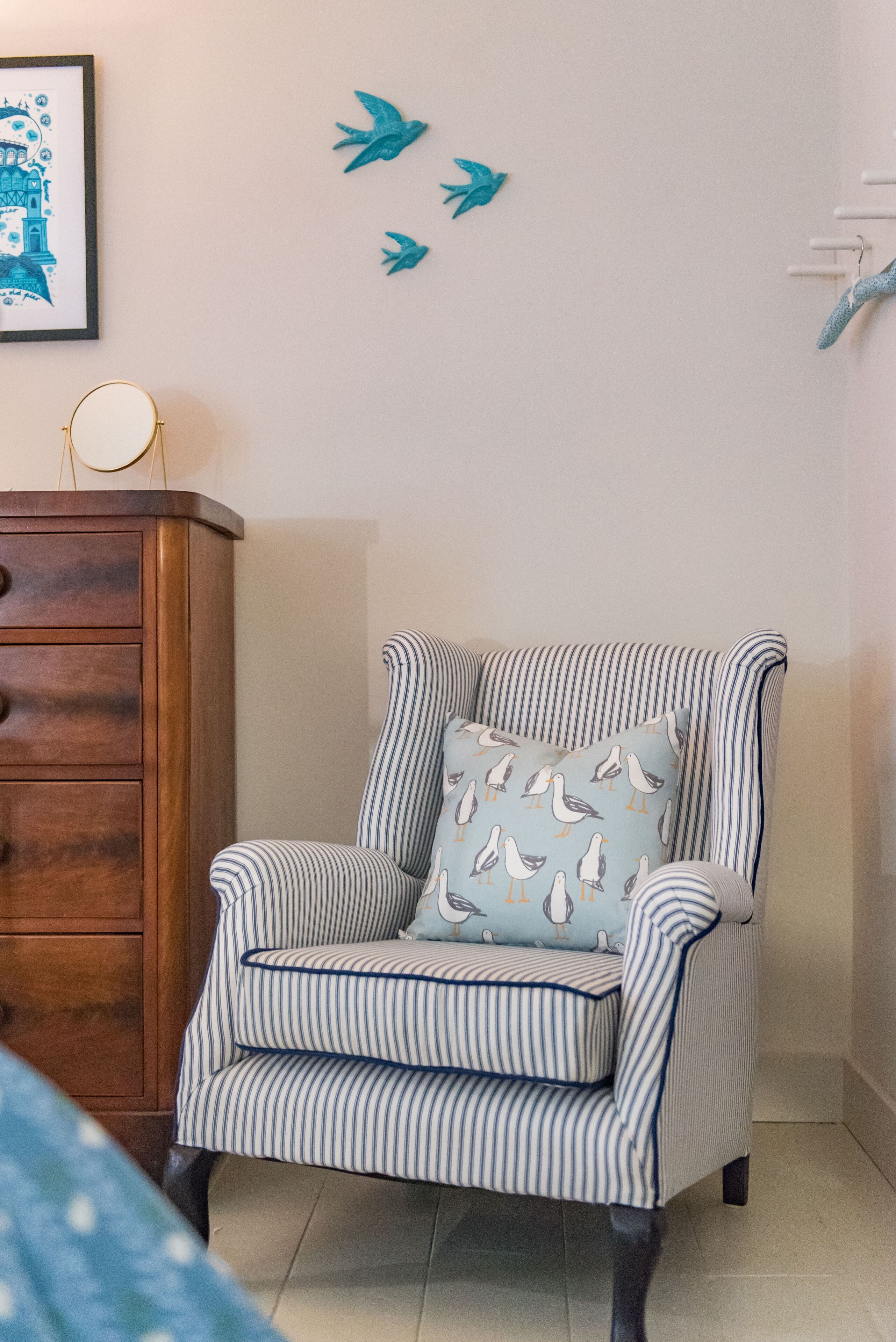 A striped armchair with a seagull pillow, a wooden dresser, and a framed picture on the wall.